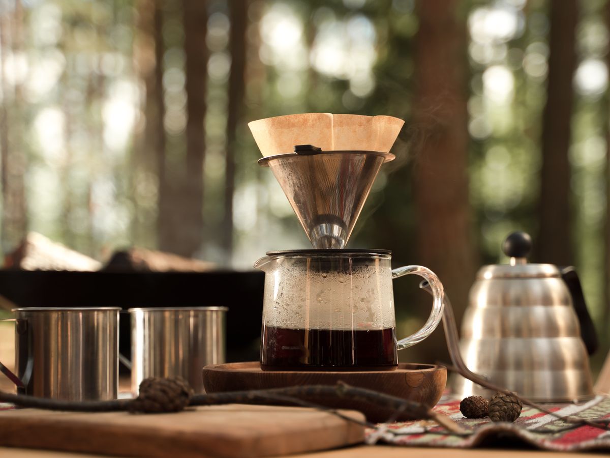 A pour-over dripper brewing coffee at a woodland campsite with steel mugs and a kettle