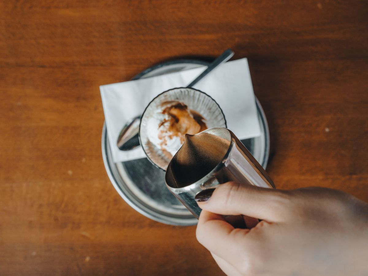 A hand pouring espresso from a pitcher over ice cream in a glass